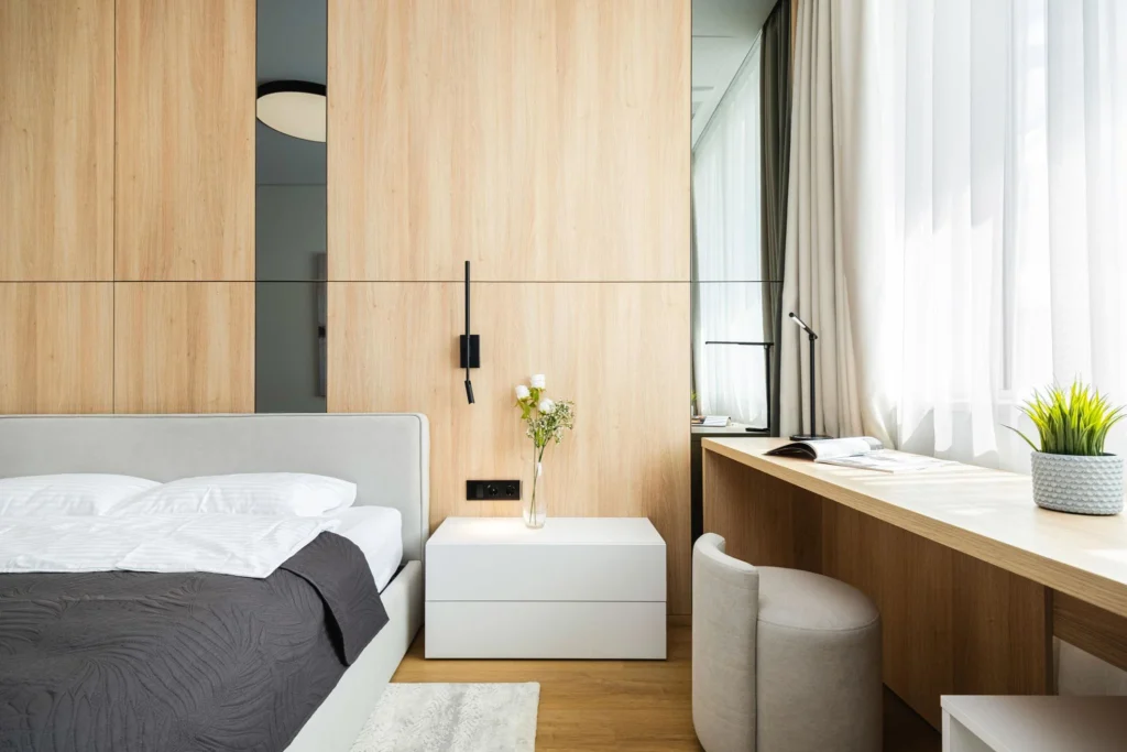 A clean minimalist bedroom with a built-in workspace, wooden panel wall, neutral bedding, modern furniture, and soft natural lighting.