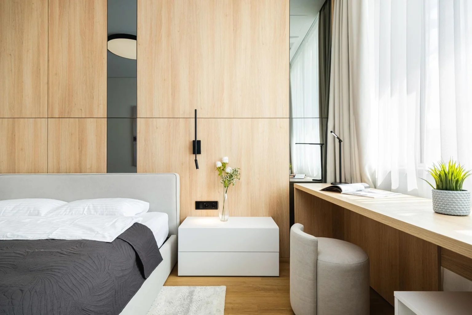 A clean minimalist bedroom with a built-in workspace, wooden panel wall, neutral bedding, modern furniture, and soft natural lighting.